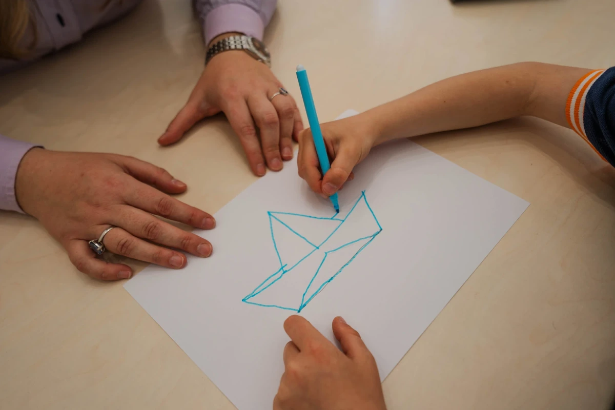 Close-up of Mariangela Allegrini's hands and a child drawing a boat with a blue felt-tip pen during a speech therapy session in Viterbo